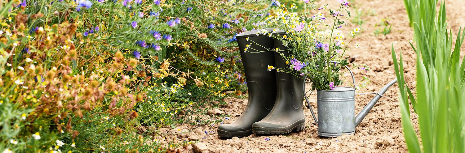 Foto: Gummistiefel und eine Grießkanne stehen auf einem Gartenweg.